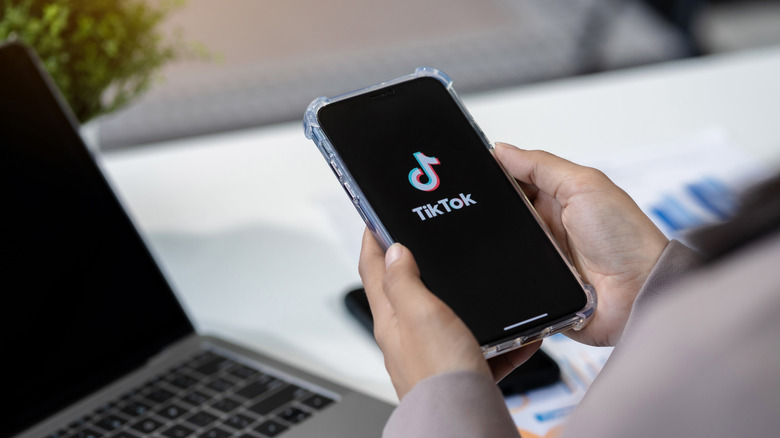 Person using TikTok on phone with computer in the background