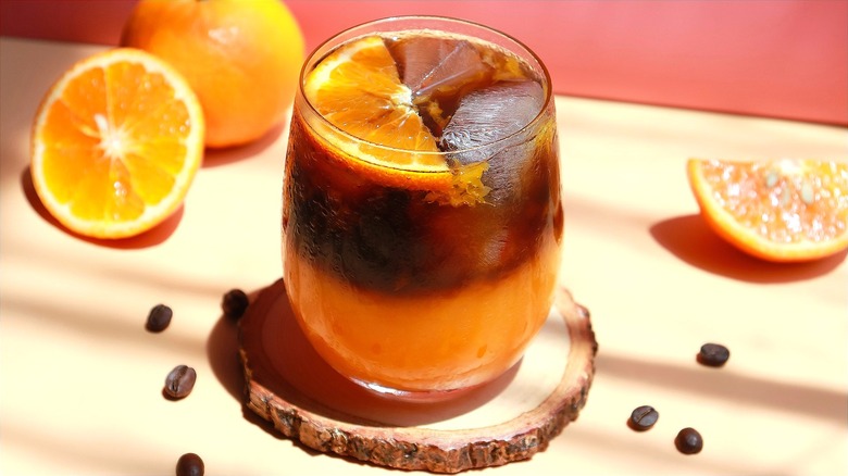 Iced orange Americano on a wooden coaster surrounded by loose coffee beans and sliced orange segments