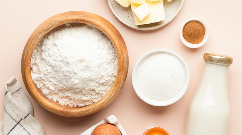 Various sized bowls of flour, butter, sugar, and cinnamon and a glass bottle of milk on a light pink background