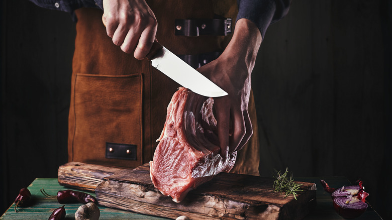 One hand holds a large knife while the other one holds a hunk of beef on a cutting board with onions, peppers, and herbs nearby