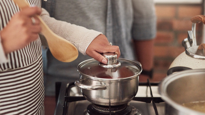 A hand grabs the lid of a closed pot sitting on the stove