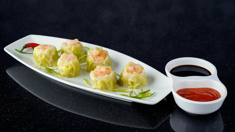 Six shumai sit on a white dish with two dipping sauces on the side.