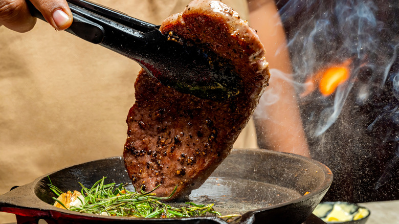 A pair of tongs lifts a steak to turn it over in a pan