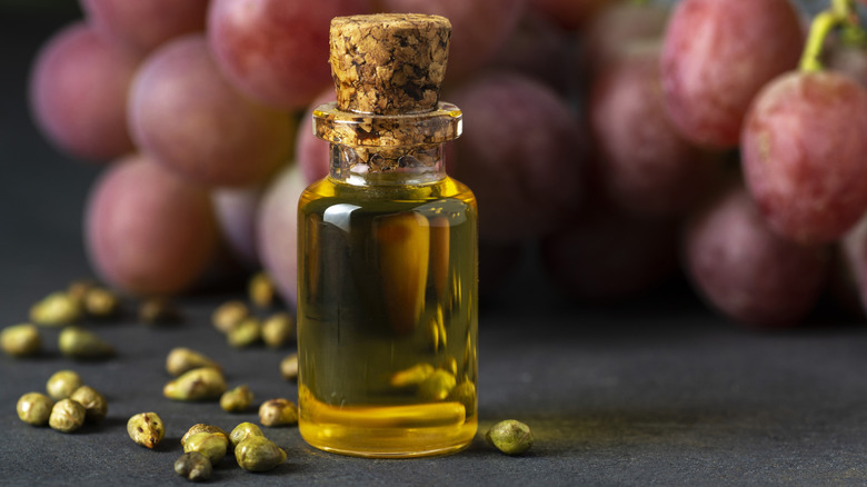A small vile of olive oil with a bunch of grapes in the background and green peppercorns on the counter