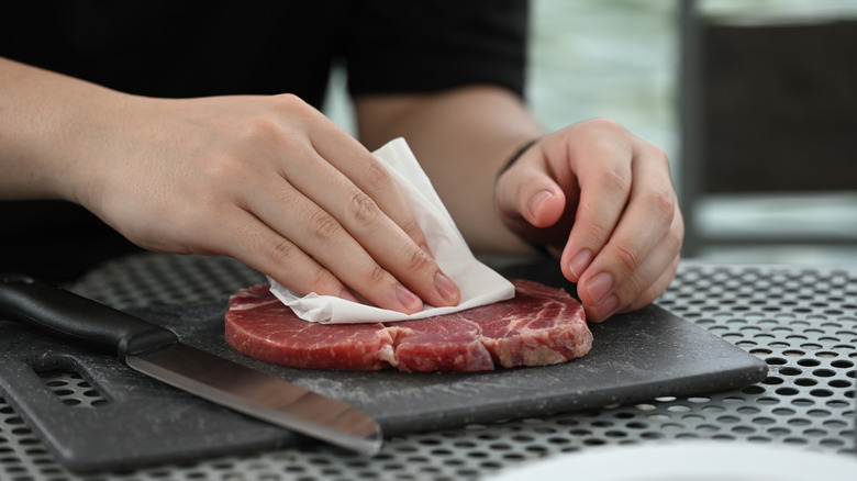 Hands use a paper towel to blot a raw cut of steak