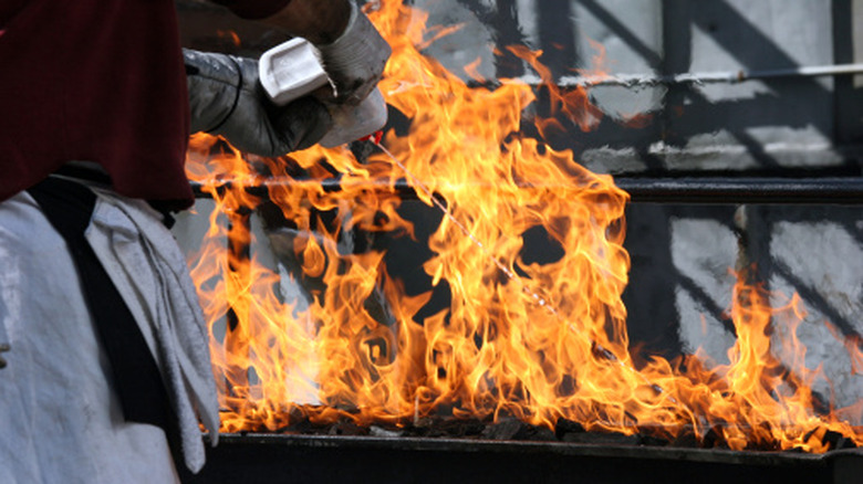 A man is squeezing a container of lighter fluid onto a charcoal grill, causing a flare up