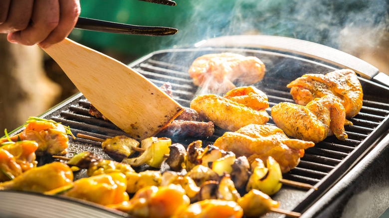 A hand turns chicken and kabobs on an electric grill with a wooden spatula