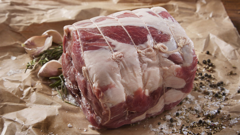 A raw pork shoulder with fat on the outside wrapped in string on a parchment paper with salt, pepper, and garlic