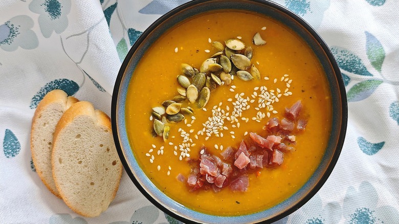 Pumpkin soup topped with bacon, sesame seeds, and pumpkin seeds next to pieces of bread.