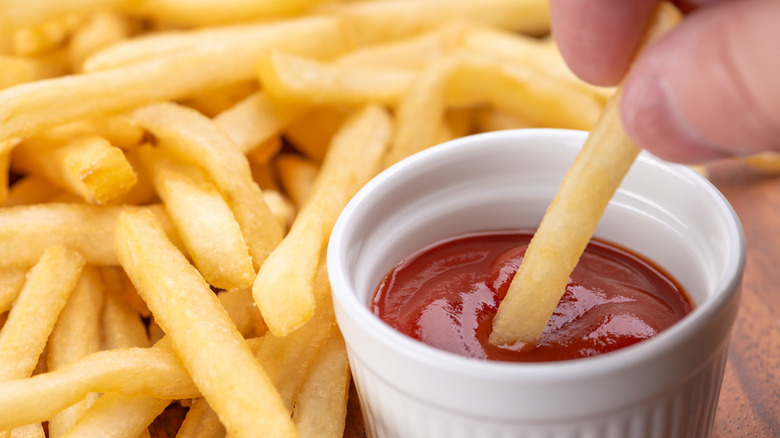 A serving of French fries with one fry being dipped in a cup of ketchup
