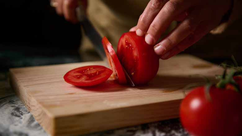 Close-up of hands slicing juicy red tomatoes on a wooden cutting board, capturing a moment in food preparation.