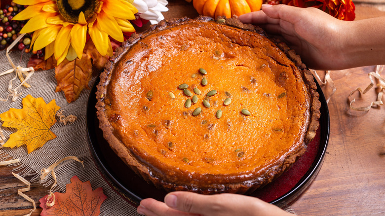 Hands holding pumpkin pie on table with autumn leaves and flowers