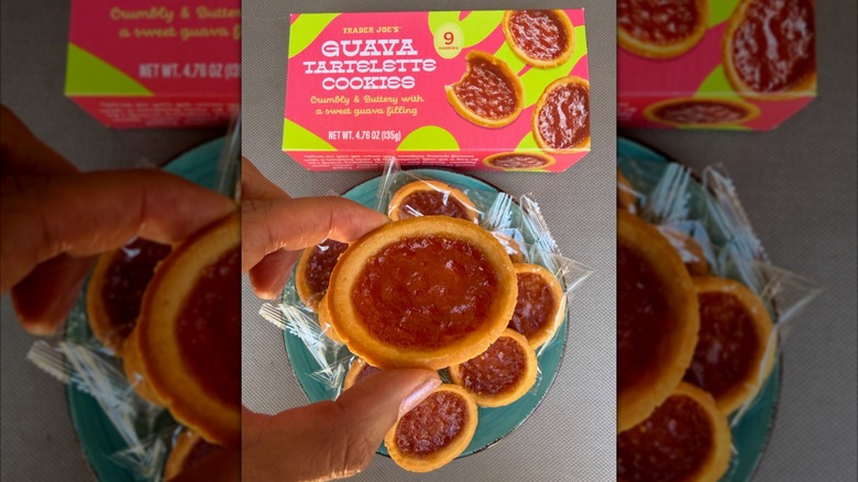 A hand holding a Trader Joe's Guava Tartelette Cookie with a box and plateful of them in background