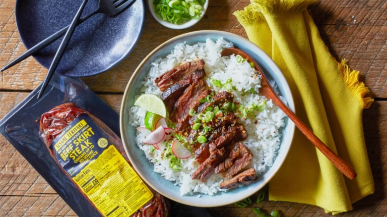 Trader Joe's Sesame Teriyaki Beef Skirt Steak next to a bowl of rice topped with sliced steak, radishes, lime wedges, and scallions