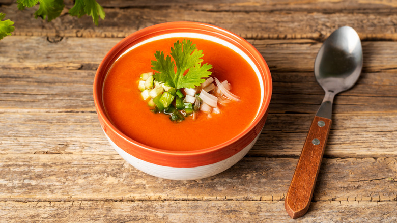 A bowl of traditional gazpacho soup garnished with fresh herbs and vegetables on a wooden tabletop.