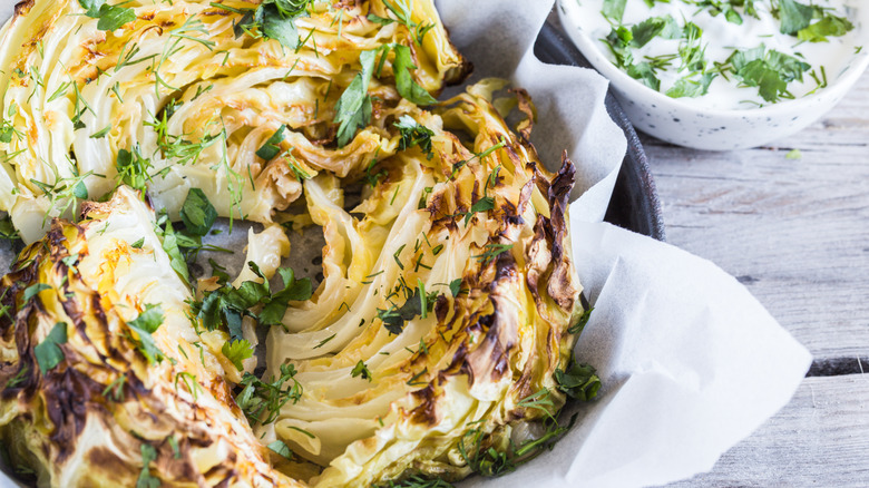 Whole roasted cabbage in a bowl, topped with herbs, with an herb sauce in the background.