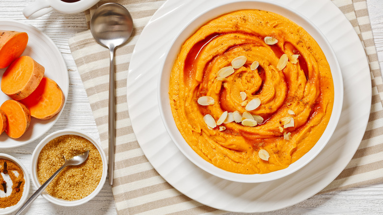 mashed sweet potatoes topped with honey and slivered almonds in a white bowl, next to a silver spoon and sliced sweet potatoes