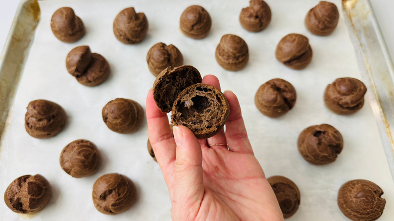 Hand holding halved chocolate pastry ball above baking sheet with chocolate pastries