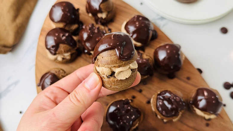 Hand holding chocolate cream puff above board with more cream puffs