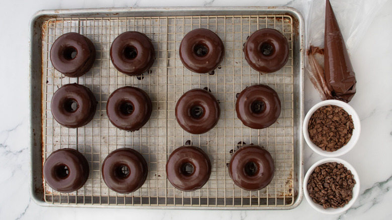 Glazed chocolate donuts on wire rack next to chocolate in piping bag and chocolate cereals in bowls