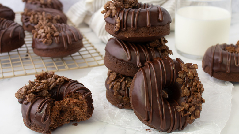 Cereal-topped chocolate donuts stacked on parchment paper with glass of milk nearby