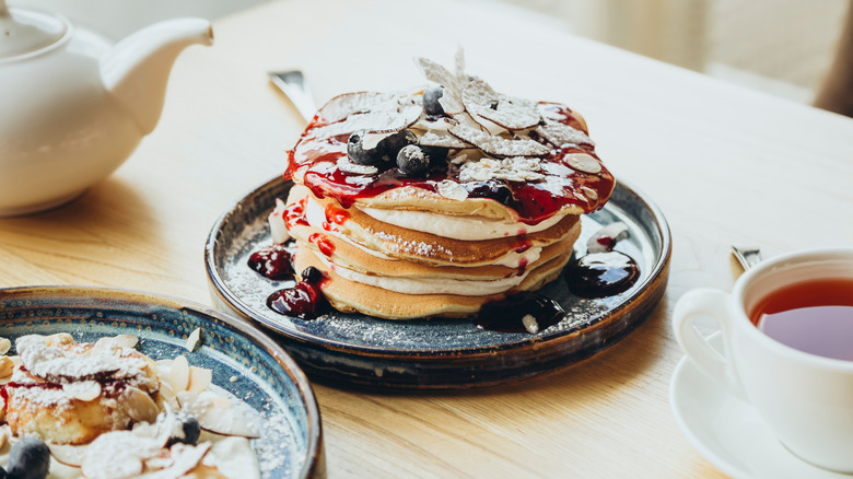 A tall stack of pancakes topped with coconut slices and blueberries, drizzled with thick jam.