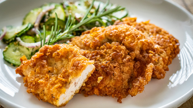 Crispy, golden brown breaded chicken cutlets on a white plate, a sprig of rosemary and a pickled cucumber salad behind.