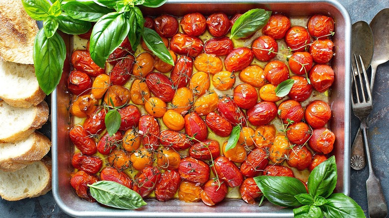 Top-down view of roasted cherry tomatoes on a silver sheet pan surrounded with fresh basil leaves and crusty bread slices.