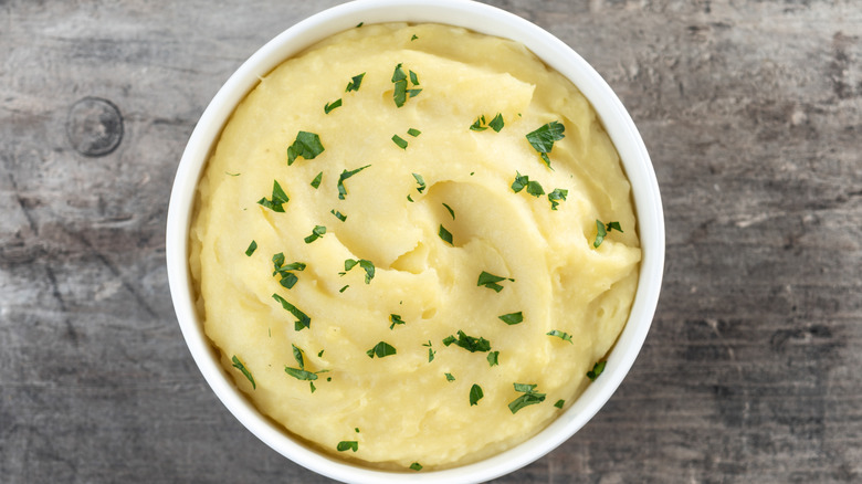 Top view of mashed potatoes in a white bowl on a wooden table sprinkled with a green herb