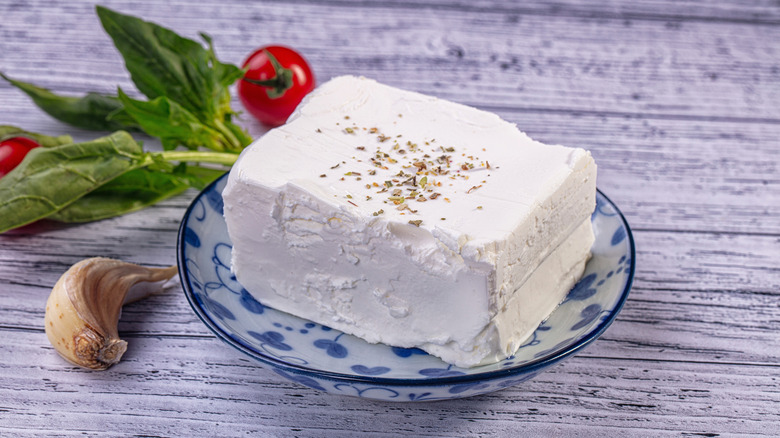 Soft white feta cheese topped with dried herbs and next to it are some basil, red cherry tomatoes and a garlic clove.
