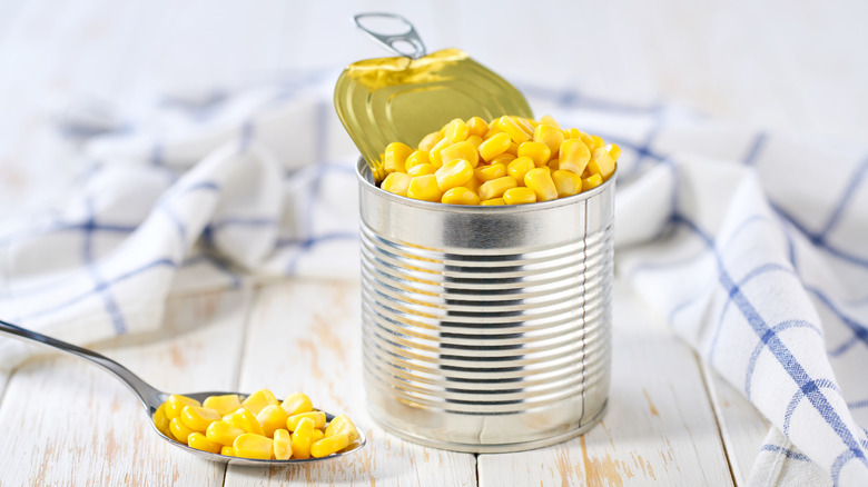 An open can of corn on a wooden surface with a napkin and spoon on the side
