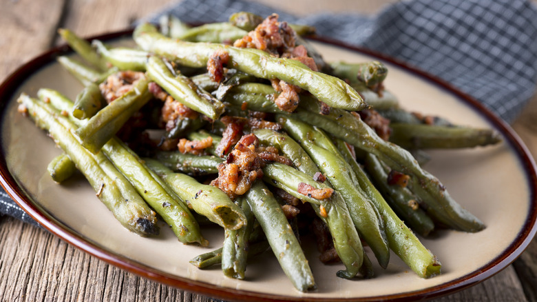 Bacon and green beans on tan plate sitting on wooden table next to napkin.