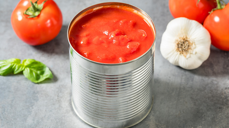 An open can of tomatoes surrounded by garlic and basil.