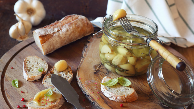Jar of garlic confit on a serving board with a partly sliced baguette