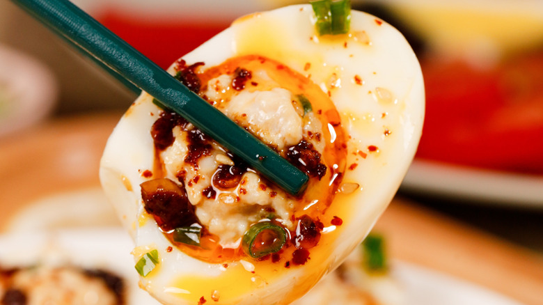 Chopsticks holding a hard-boiled egg with chili oil, scallions, and a dumpling in the middle
