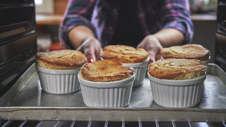 Hands reach into an oven to retrieve a sheet of golden soufflés in ramekins.