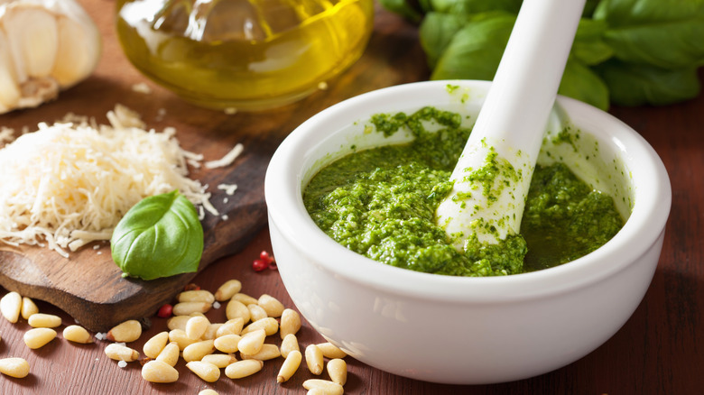 A white bowl of pesto is surrounded by ingredients like parm and pine nuts.