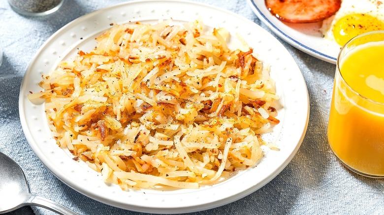 Cooked, shredded hash browns on a white plate next to a plate of eggs and Canadian bacon and a small glass of orange juice