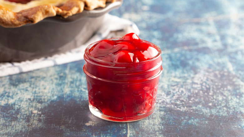 Glass jar of canned cherry pie filling with a pie in the background