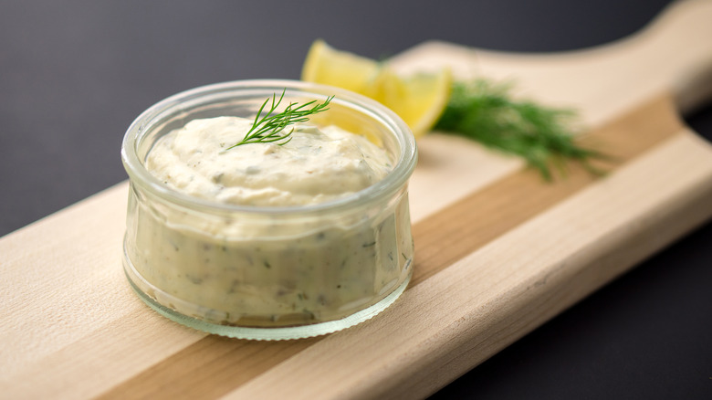 Homemade tartar sauce made of fresh mayonnaise, lemon and various herbs, served in a glass dipping bowl on a wooden serving board.