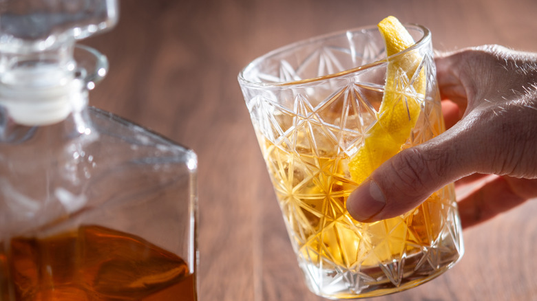 A hand holding a rusty nail cocktail garnished with a lemon twist