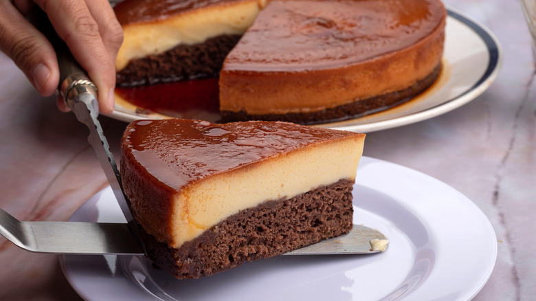Hands holding a knife and spatula, placing a slice of chocoflan on a plate while the rest of the chocoflan sits behind it on another plate