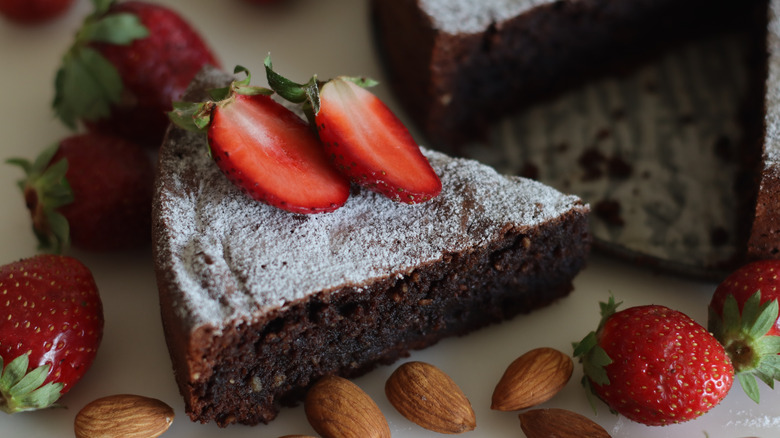 A slice of torta caprese, topped with powdered sugar and two strawberry halves, surrounded by strawberries and almonds