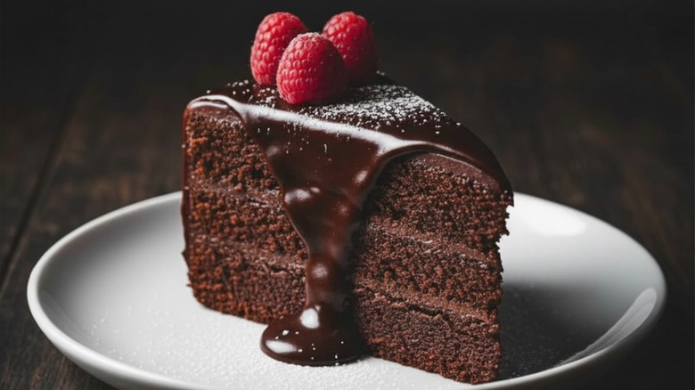 A slice of chocolate cake with three layers, a melty chocolate frosting, three raspberries on top, and powdered sugar sits on a white plate
