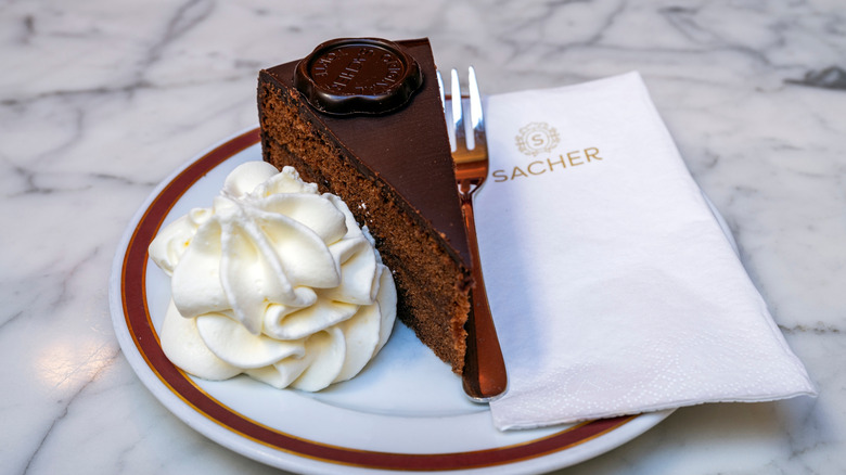 A slice of Sachertorte on a plate with a pile of whipped cream, next to a fork and a white napkin with the Sacher name and logo