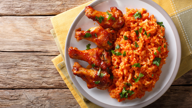 Four fried chicken pieces covered with red sauce on a white plate next to jollof rice