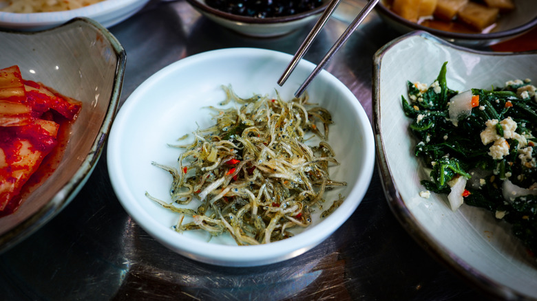 chopsticks pointing to a side of myeolchi bokkeum next an array of other banchan