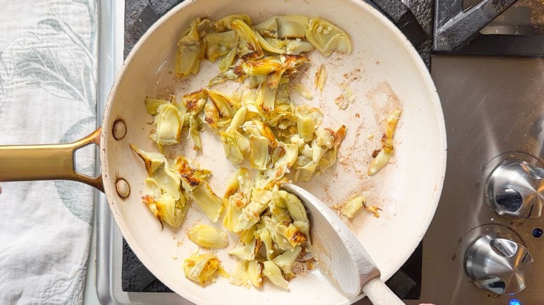 Stirring browned artichoke heart quarters and garlic in skillet on stovetop