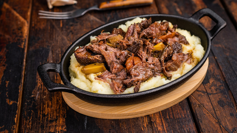 Braised beef cheeks in a stew sit atop a bed of mashed potatoes, served in a skillet