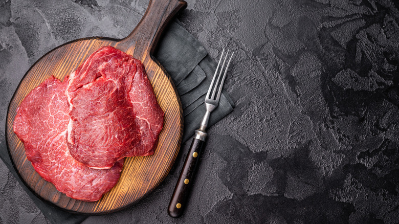 Beef cheeks sit atop a wooden cutting board and next to a piercing fork